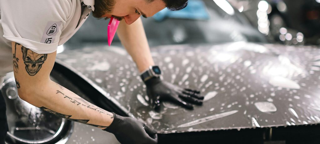 A tattooed man is washing a car, focused on cleaning the vehicle's surface with a cloth