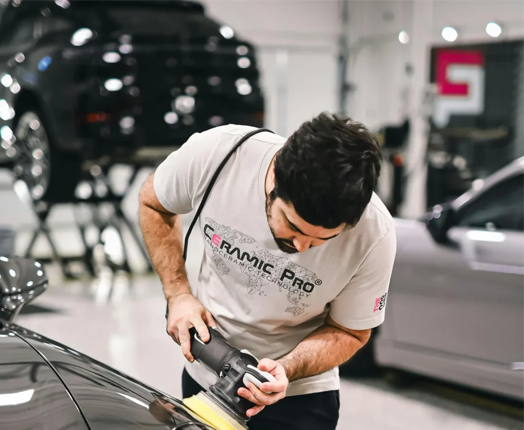 A man operates a car wash machine, cleaning a vehicle with soap and water in a bright, sunny environment