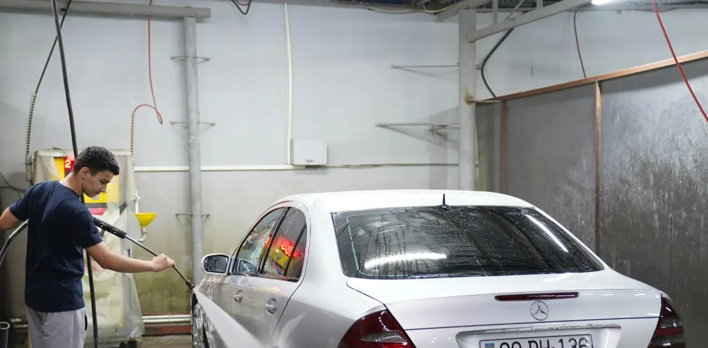A man scrubs a car with soap in a car wash, surrounded by water sprays and cleaning equipment