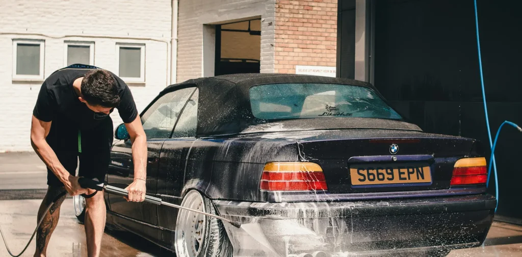 A man scrubbing a car with a sponge, water splashing around him in a sunny outdoor setting