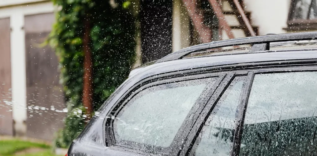 A man washes his car with a hose, spraying water over the vehicle in a sunny outdoor setting