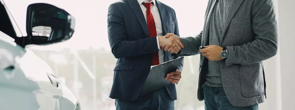 Two men shaking hands in front of a parked car, symbolizing a business agreement or partnership