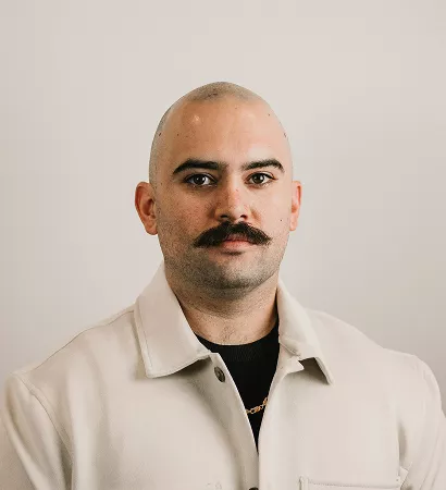 A man with a moustache wearing a white jacket, standing confidently with a neutral expression