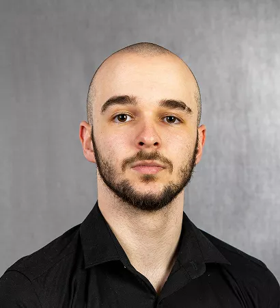 A man with a beard wearing a black shirt, looking directly at the camera with a neutral expression