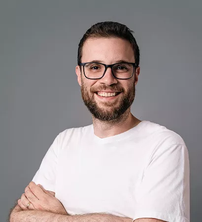A man with glasses and a beard wearing a white t-shirt, looking directly at the camera with a neutral expression