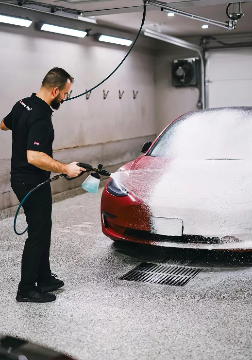 A man washes a red Tesla car inside a garage, surrounded by cleaning supplies and tools